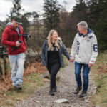 A Lake District wedding videographer walks alongside a couple holding hands on a scenic forest path near a lake. The videographer, wearing a red jacket and green cap, captures the couple’s joyful expressions in this peaceful, natural setting.